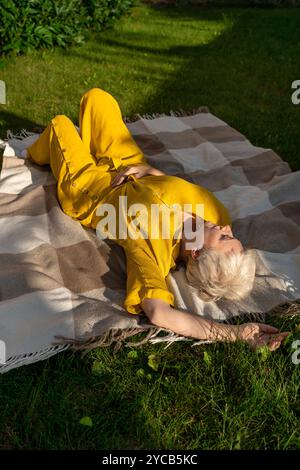 A mature woman with white hair, dressed in a vibrant yellow outfit, relaxes on a plaid blanket in a lush garden. Her eyes are closed, evoking a sense Stock Photo