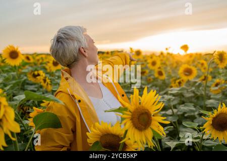 A mature woman with short grey hair is seen enjoying a serene sunset amidst a vibrant sunflower field. She wears a bright yellow jacket, complementing Stock Photo