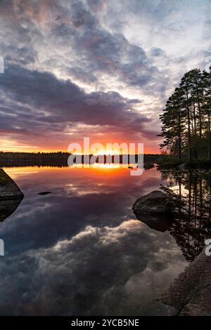A sunset at Tiveden National Park casts vibrant hues over a serene lake, reflecting the sky and silhouetted trees in Sweden Stock Photo