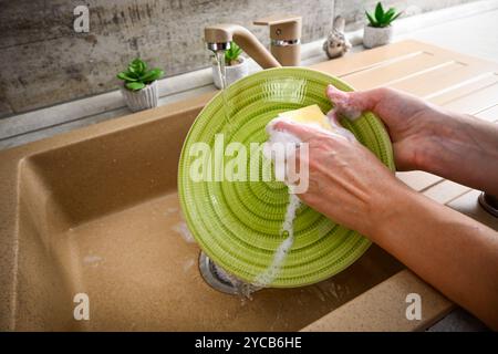 Hands washing a green plate with a yellow sponge under running water in a kitchen sink. Stock Photo