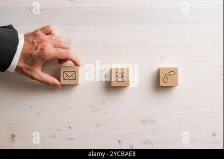 A hand in a suit arranges wooden blocks with icons representing email, group communication, and phone contact on a light wooden surface, symbolizing d Stock Photo