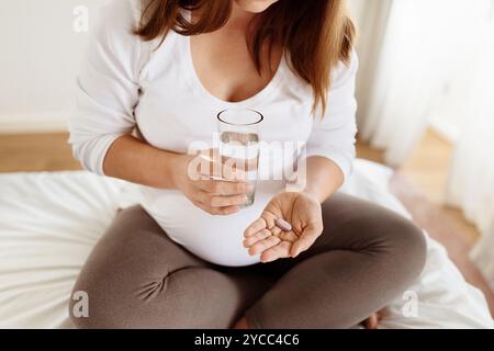 Pregnant woman sitting on bed taking prenatal vitamins with water Stock Photo