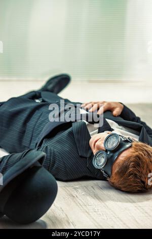 Tired businessman in a suit and tie lies on the floor. Wearing a hat and steampunk goggles. Looking defeated after a long day at work. Failure. And de Stock Photo