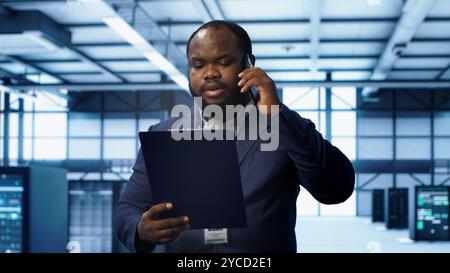 IT professional looking over data center checkup checklist paperwork, talking on phone in server farm. African american man surrounded by server rigs, chatting during telephone call, camera A Stock Photo
