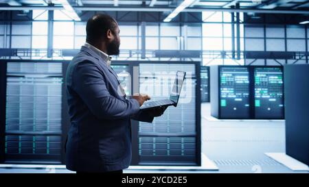 African american man walking in high tech facility, using laptop to patch network issues leading to slow data transfer rates. Data center worker fixing errors affecting supercomputers, camera B Stock Photo