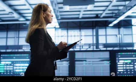 Engineer in server hub checking recovery plan on tablet, monitoring rackmounts energy consumption. Woman in data center making sure rigs are functioning optimally, doing routine disk checks, camera A Stock Photo