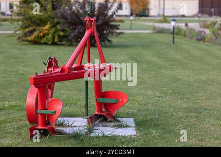 Red agricultural plow standing on a grassy field near a farm on a sunny day. Concept of farming equipment, traditional agriculture and rural lifestyle Stock Photo