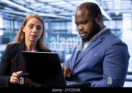 Teamworking admins in server room using laptop to implement and maintain security measures, protecting systems from unauthorized access. Data center IT professionals colleagues doing hardware upgrades Stock Photo