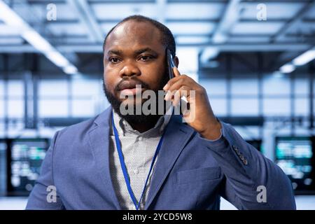 Team leader overseeing operations in modern data center, coordinating tasks during phone call. Close up of IT professional in server farm managing systems and communicating via smartphone Stock Photo