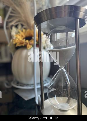 Elegant hourglass measures time with sand trickling through glass bulbs. In background, a blurred vase with yellow flowers adds warmth to the scene. Stock Photo