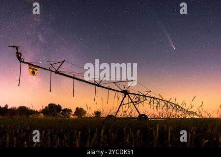 Comet Tsuchinshan-Atlas above a field irrigator with the Milky Way to the left. A farmhouse with illuminated windows is in the background. Sunset Stock Photo