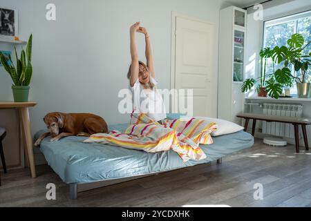 Positively woke up in morning, yawning woman stretched sits on bed with dog optimistic started day. Stock Photo