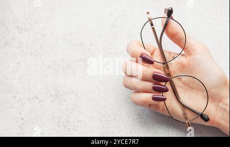 A hand with long burgundy nails gracefully holds a pair of round glasses above a light textured surface, illuminated by soft natural light. Stock Photo