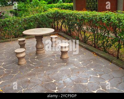 A stone table with matching stools stands on a patterned stone floor, surrounded by lush garden greenery. Stock Photo