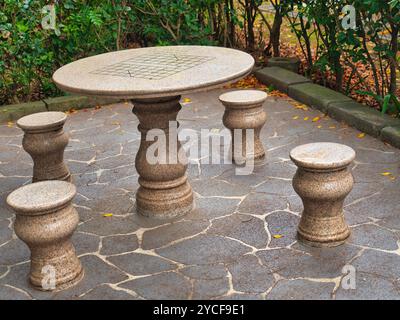 A stone table with matching stools stands on a patterned stone floor, surrounded by lush garden greenery. Stock Photo