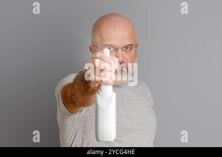 Bald bearded man holds spray bottle, sprays chemicals. Gray background. Disinfection of premises Stock Photo