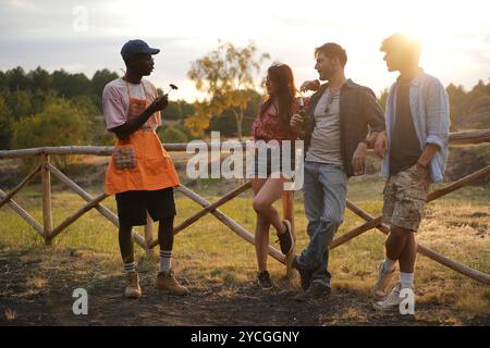 Multiracial group of young friends hanging out at rustic fence during sunset. Natural lifestyle moment of diverse people enjoying summer evening toget Stock Photo