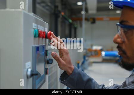 African American engineer, wearing a gray work coat and a blue helmet, controlling production and programming a machine in a factory. Stock Photo