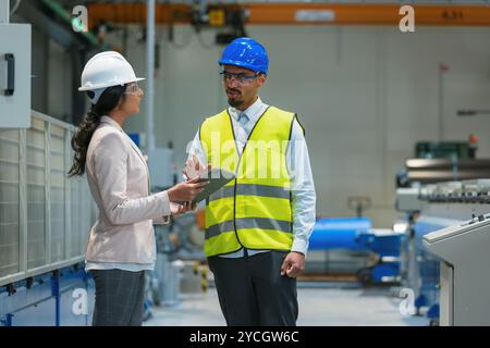 Worker consulting with a woman manager in the factory, analyzing graphs and data on a tablet to improve production processes. Stock Photo