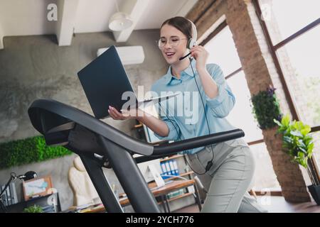 Photo of happy positive lady recruiter dressed shirt headphones running treadmill speaking modern gadget indoors workplace workstation loft Stock Photo