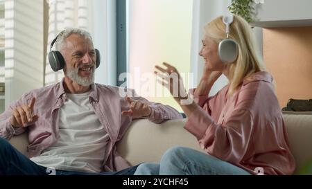 Joyful senior elderly Caucasian married couple grandparents man woman listening music using headphones dancing fooling around having fun carefree Stock Photo