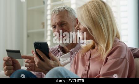 Elderly happy Caucasian married couple senior man woman retired make order purchase at online shop using phone pension credit card enjoy easy buying Stock Photo