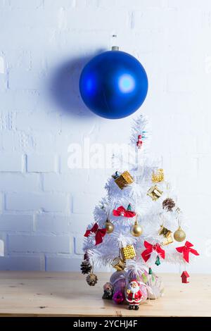 Small white Christmas tree decorated with golden and red ornaments, pine cones, and miniature Santa figurines on a wooden table against a white brick Stock Photo