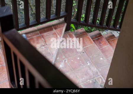 A view of a staircase with terracotta tiles, surrounded by wooden railings. The stairs descend into a green outdoor area, with some foliage visible in Stock Photo