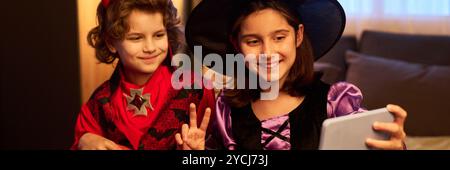 Header of happy youthful girl and boy in Halloween costumes taking selfie while sitting on bed in front of camera at home Stock Photo