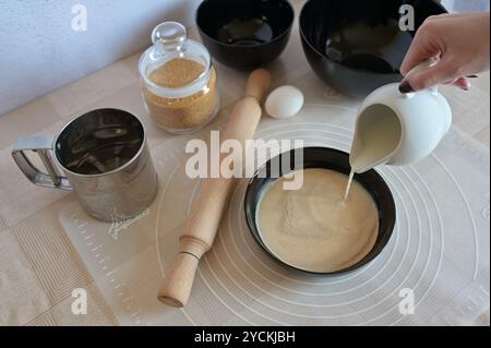Baking ingredients for baking sweet buns, yeast in black bowl dishes, creamer with milk in a woman hand, rolling pin on a table Stock Photo