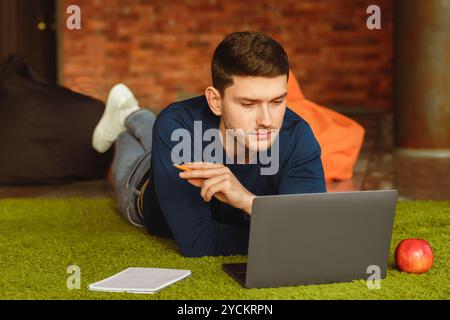 Millennial Guy Using Laptop Lying On Floor At Home Stock Photo