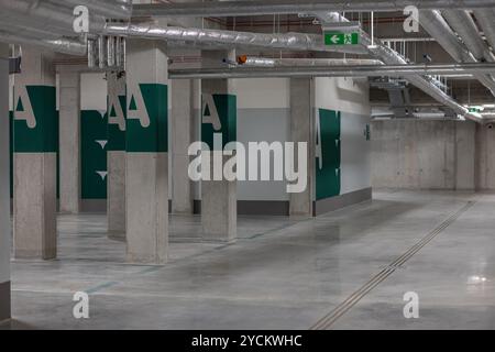 Empty underground parking garage with concrete pillars and ventilation pipes, marked with green letters and emergency exit signs Stock Photo