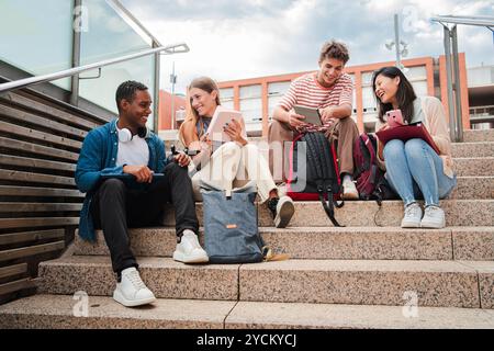 Group of diverse young students talking and smiling while sitting on university campus stairs. Multiracial friends enjoying casual conversation, holding tablets and smartphones in a relaxed academic. High quality photo Stock Photo
