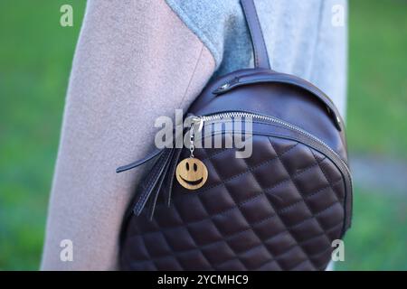 Woman with wooden keychain in shape of smiley face and black leather decor on her backpack outdoors, closeup Stock Photo