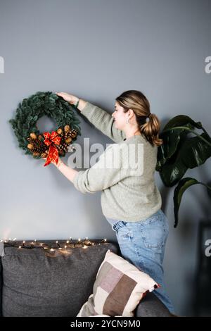 Woman hanging handmade Christmas wreath with pinecones and red ribbon. DIY holiday decor adds a personal touch to festive home decorating. Stock Photo
