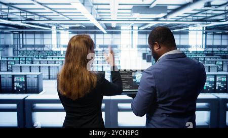 Teamworking programmers in data center overseeing server operations using laptop and tablet. Coworkers ensuring optimal performance and data integrity in server room using devices, camera B Stock Photo