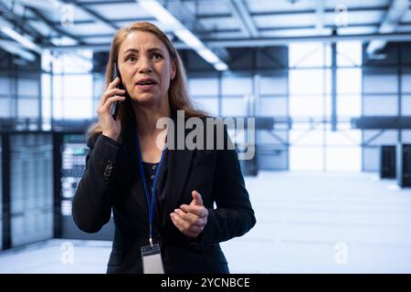 Businesswoman speaking on phone in high tech data center housing advanced computing equipment. IT expert delegating tasks in technologically advanced environment during telephone call Stock Photo