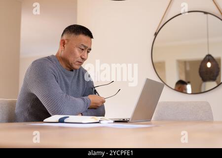Asian senior man working on laptop at home, holding glasses, focused on screen Stock Photo