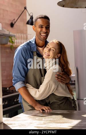 multiracial couple hugging in kitchen, wearing aprons, smiling joyfully together, at home Stock Photo