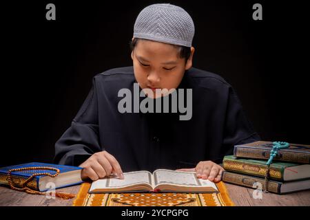 A little Asian Muslim boy is sitting and reading the Quran at the wooden table at his house during the night during the month of Ramadan, Arabic calli Stock Photo