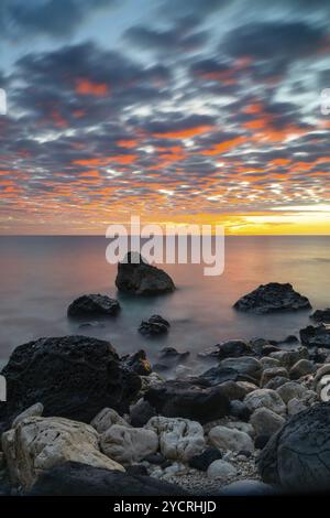 A colorful sunrise at Cala Gonone with black and white rocks and boulders in the foreground Stock Photo