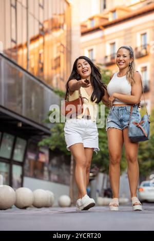 Two young women are walking down a city street, one of them is pointing at something and they are both smiling Stock Photo