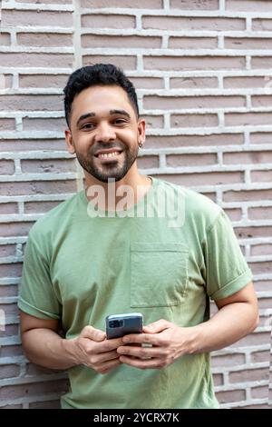 Young man is smiling while using a smartphone and leaning against a brick wall Stock Photo