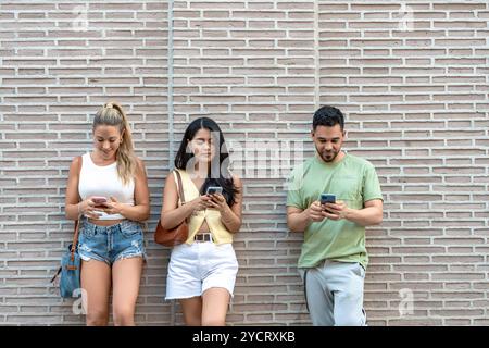 Three friends standing against a brick wall in the city, engrossed in smartphones, browsing, scrolling, and texting, showcasing the modern lifestyle of constant digital connection Stock Photo