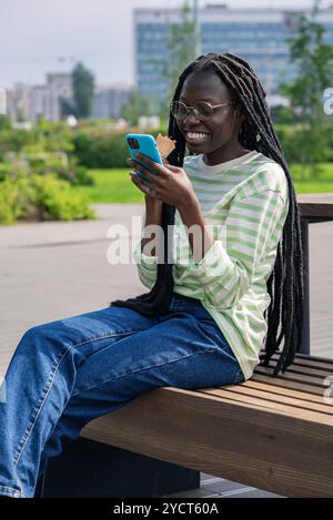 Young woman is sitting on a bench, enjoying an ice cream cone and scrolling on her smartphone. She is happy and enjoying her break Stock Photo