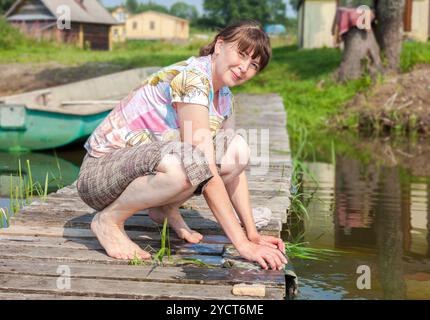 Young woman washes clothes on wooden jetty by the lake in summer sunny day Stock Photo