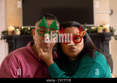 Chrsitmas time, at home, multiracial senior couple wearing festive glasses making funny faces Stock Photo