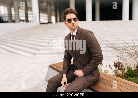 Portrait of stylish photogenic young businessman in brown suit and sunglasses sitting confidently on city bench, exuding professionalism and success Stock Photo