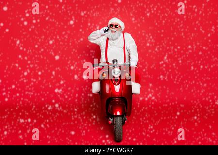 Portrait of nice attractive bearded cheerful funny Santa wearing cap hat riding motor bike talking on phone taking orders isolated over bright vivid Stock Photo