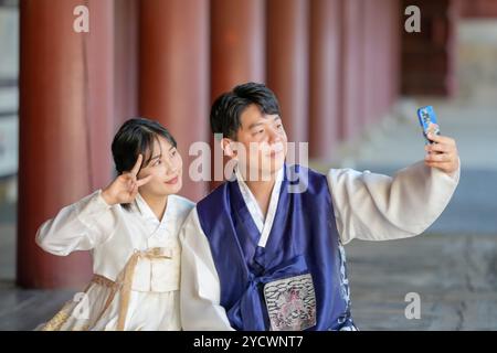 A Korean couple, a man in his 30s and a woman in her 20s, are happily wearing Hanbok and taking a selfie with a smartphone inside a historical buildin Stock Photo